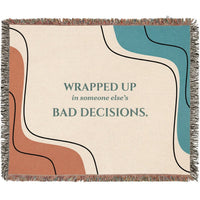 A woven cotton throw blanket with fringe edges, featuring an abstract design with wavy lines in terracotta, teal, cream, and black, with the text 'WRAPPED UP in someone else's BAD DECISIONS.' printed in teal in the center..