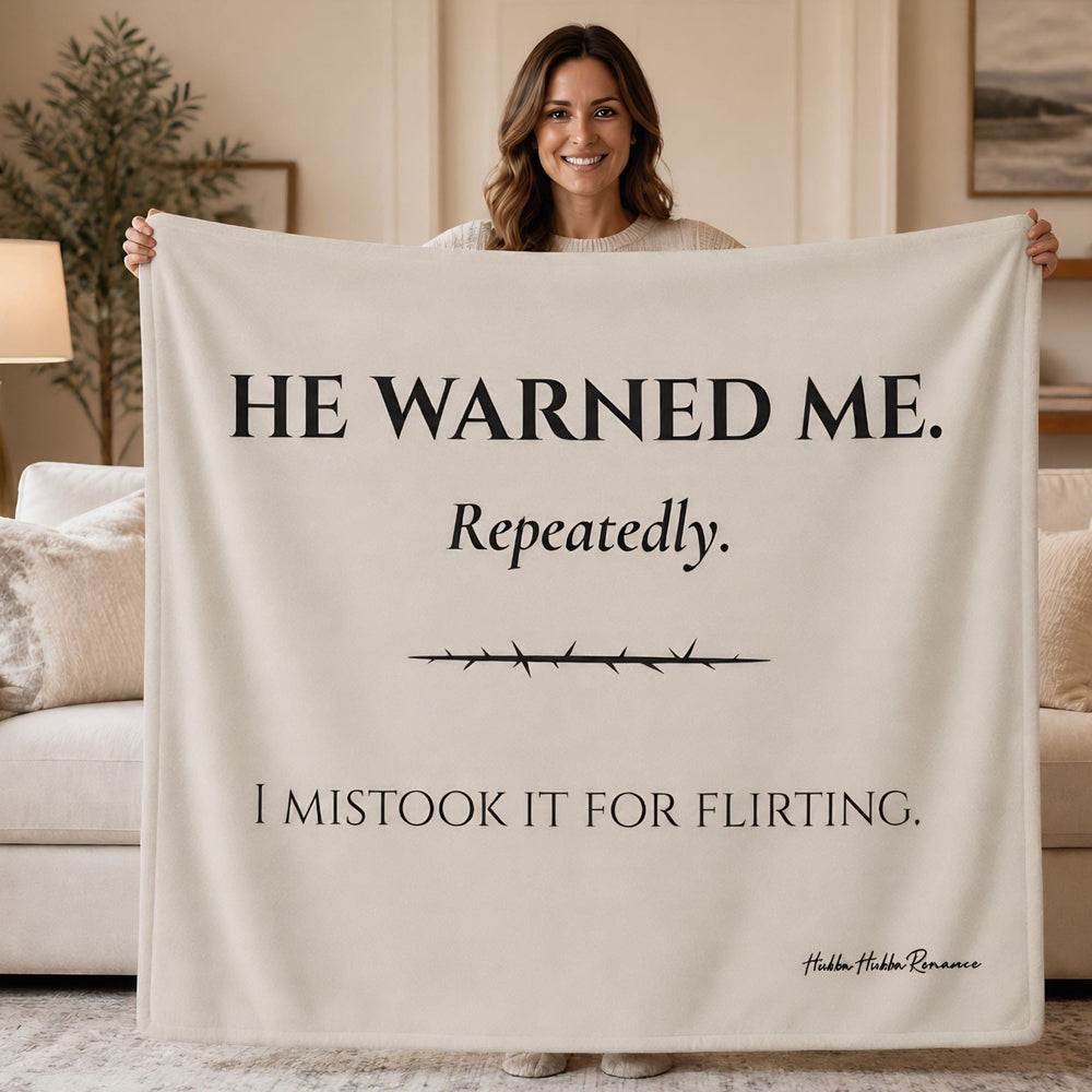 A woman stands in a living room holding up a large cream-colored blanket or throw that reads, 'HE WARNED ME. Repeatedly. I mistook it for flirting.