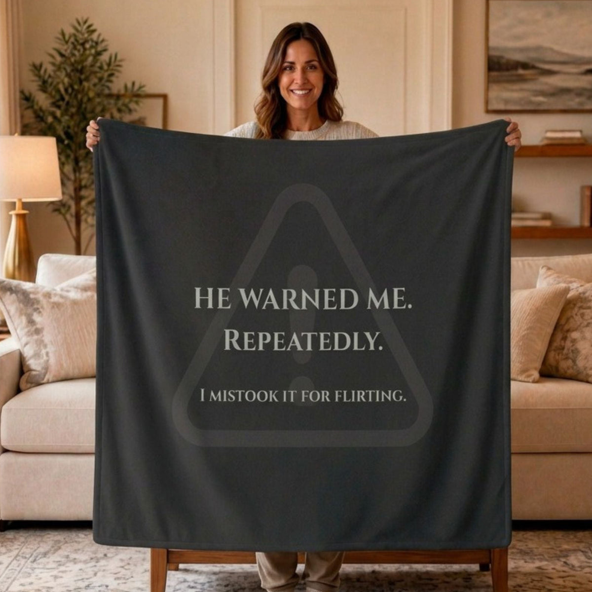 A woman stands in a living room holding up a large dark charcoal gray sweatshirt blanket or throw with creamtext that reads, 'HE WARNED ME. REPEATEDLY. I mistook it for flirting. There is a very faded warning symbol graphic that sits behind the text.