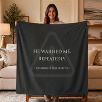 A woman stands in a living room holding up a large dark charcoal gray sweatshirt blanket or throw with creamtext that reads, 'HE WARNED ME. REPEATEDLY. I mistook it for flirting. There is a very faded warning symbol graphic that sits behind the text.