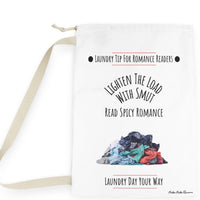 This laundry bag features a white background with a colorful pile of clothes  at the bottom. Above the pile, black text reads: “Lighten the Load with Smut” in an arched layout. Below that, in a straight line, it says: “Read Spicy Romance.” At the top, the header “Laundry Tip for Romance Readers” is centered between two black dots and underlined in red. The bottom of the image reads: “Laundry Day Your Way” in black text, underlined in red. The bag includes a drawstring closure and a canvas carry strap.