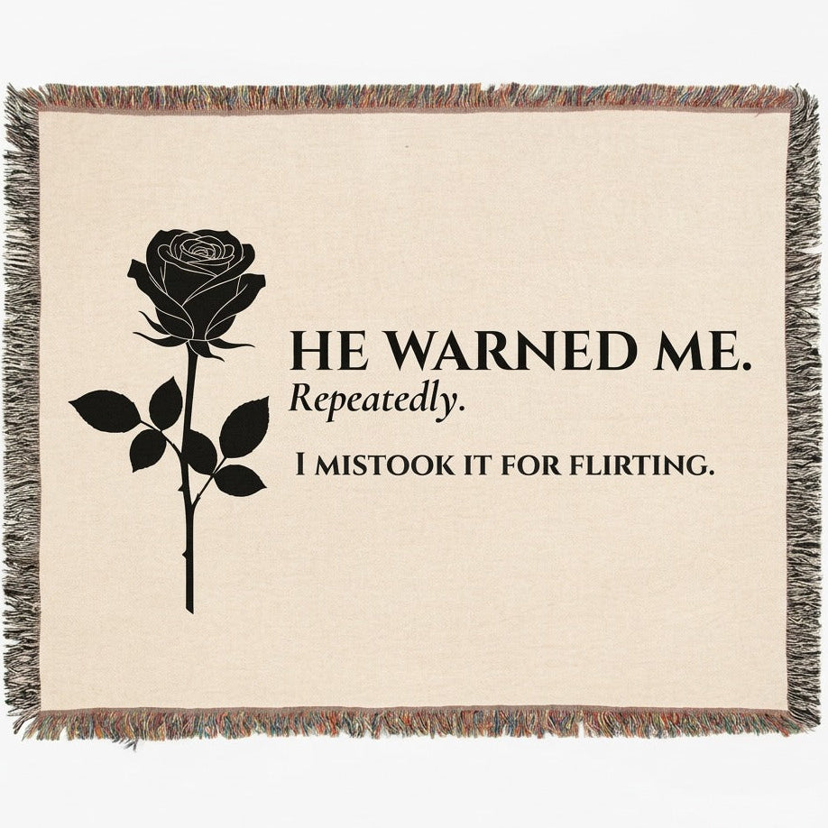 A woven cotton throw blanket with fringed edges, featuring a black rose graphic and the text 'HE WARNED ME. Repeatedly. I mistook it for flirting.