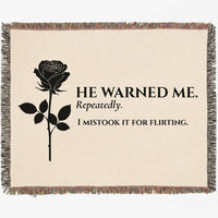 A woven cotton throw blanket with fringed edges, featuring a black rose graphic and the text 'HE WARNED ME. Repeatedly. I mistook it for flirting.