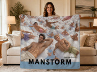A smiling woman stands in a cozy living room holding the Manstorm sweatshirt blanket. The blanket features a humorous romance-reader design showing dozens of handsome, muscular book-boyfriend style men floating among fluffy white clouds in a pale blue sky. Two larger figures dominate the foreground while smaller men drift throughout the background, creating a playful fantasy weather scene. The word "Manstorm" appears prominently across the bottom of the blanket.