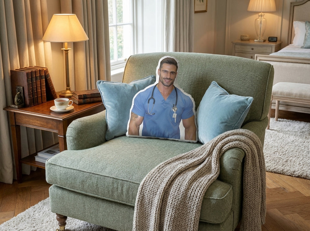 Cozy bedroom reading nook with a green upholstered armchair and a chunky knit throw blanket draped over the arm. A custom-shaped book boyfriend pillow featuring a handsome man in blue medical scrubs with a stethoscope is propped against blue accent pillows. A side table with stacked books and a lit lamp sits beside the chair, with a bed visible in the background.