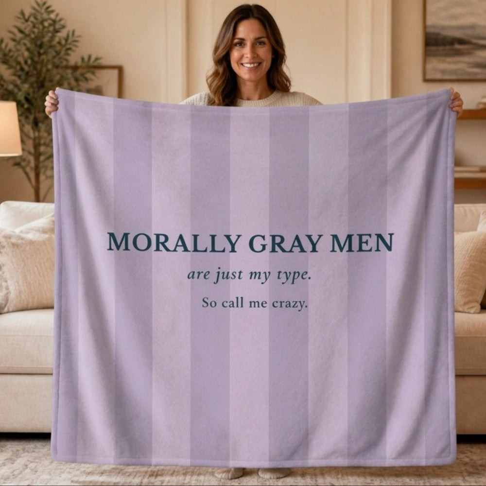 A woman isstanding and holding a  lavender and light gray striped blanket in a living room setting. The blanket features the text 'MORALLY GRAY MEN are just my type. So call me crazy.