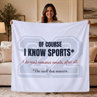 A woman stands in a living room holding up a large light grayish blue sweatshirt  blanket  that reads, 'OF COURSE I KNOW SPORTS* I do read romance novels, after all. *The stuff that matters.