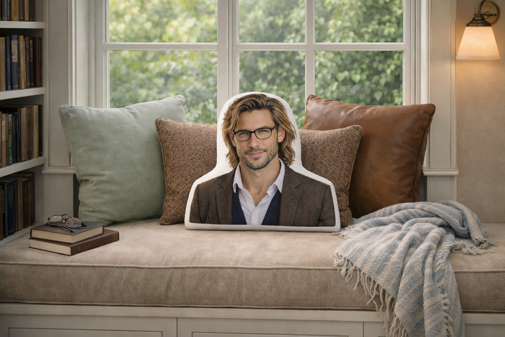The Professor academic romance book boyfriend pillow styled in a built-in reading nook with a large window behind the seating area, a light khaki cushion, and a row of decorative pillows including sage green, brown tweed, and leather. A light blue striped cozy blanket and stacked books complete the calm, bookish setting designed for readers who love intelligent, slow-burn romances.