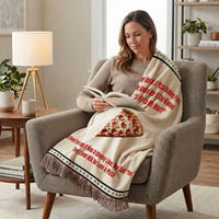 A woman sits in an armchair reading a book while wrapped in a cream throw blanket printed with red humerous pizza-themed text and a slice of pepperoni pizza graphic. 