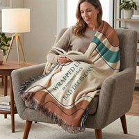 A woman sits in an armchair reading a book, wrapped in a cream throw blanket with brown, teal, and black stripes and the phrase 'WRAPPED UP in someone else's BAD DECISIONS.'.