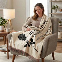 A woman sits in an armchair reading a book, wrapped in a cream throw blanket printed with a black rose and the phrase 'HE WANTED ME. Repeatedly. I mistook it for flirting.