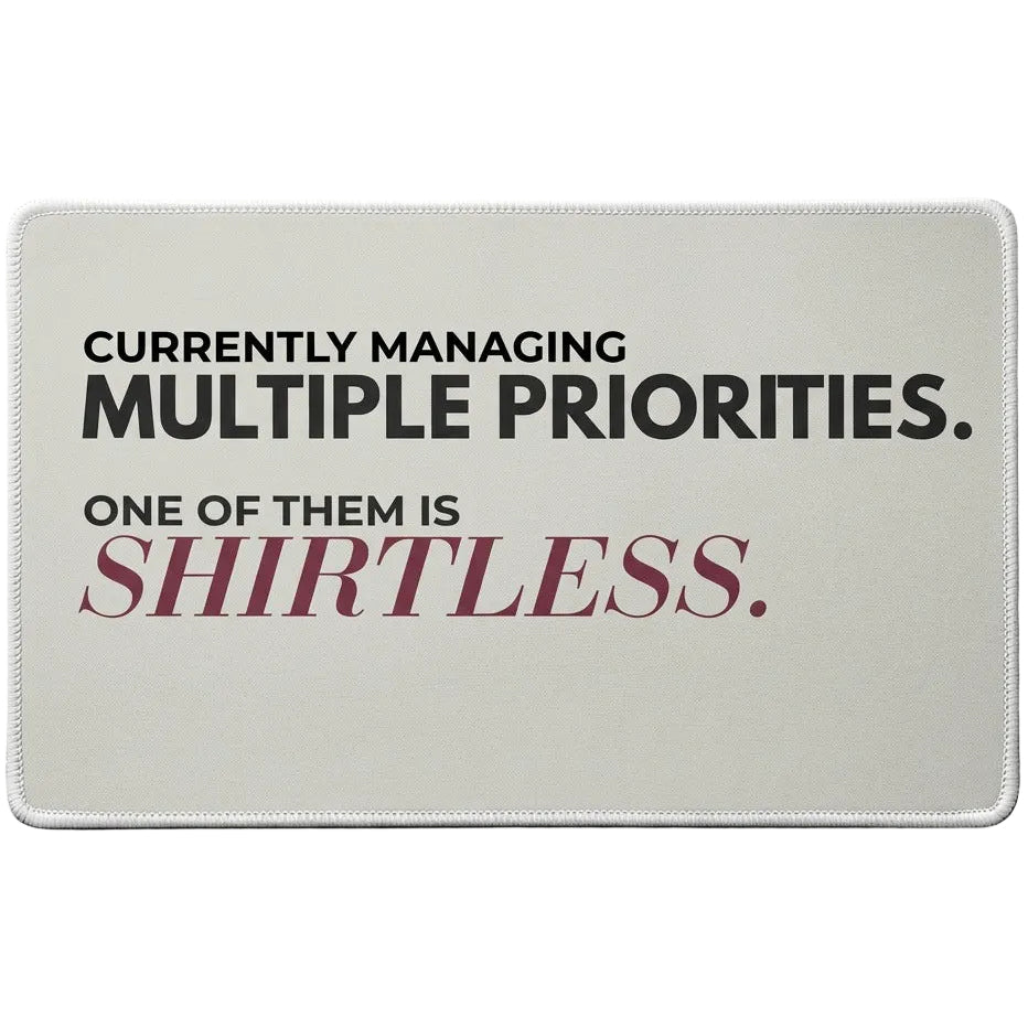 A rectangular sage green desk mat with rounded corners featuring the text 'CURRENTLY MANAGING MULTIPLE PRIORITIES. ONE OF THEM IS SHIRTLESS.