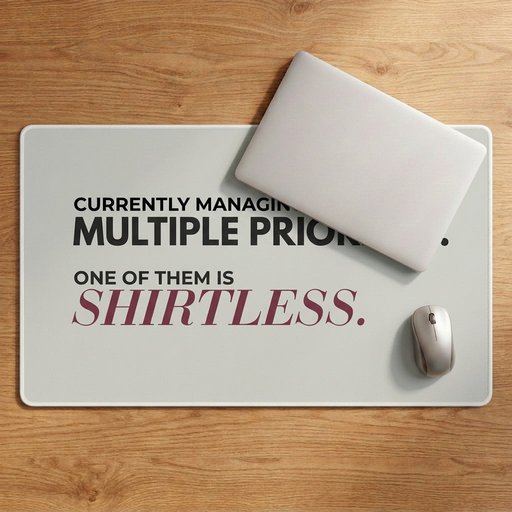 A rectangular desk mat with humorous text on a wooden desk, styled with a closed silver laptop and wireless mouse.The text reads 'CURRENTLY MANAGING MULTIPLE PRIORITIES. One of them is shirtless.