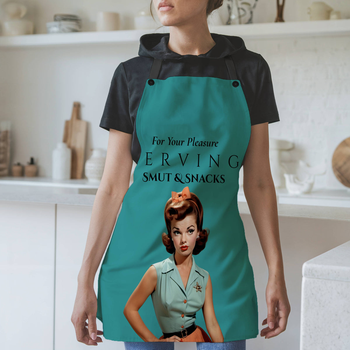 A woman wearing a teal apron with a retro pin-up girl graphic and the text 'For Your Pleasure. SERVING SMUT & SNACKS' over a black t-shirt, standing in a bright kitchen setting.